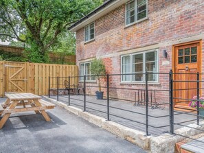 A brick building with a wooden door, a fenced patio with a table and chairs, and a large tree.