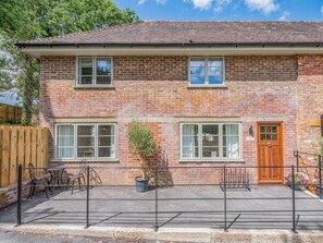 A two-story brick house with a front door, two windows on the ground floor, and a small patio area with a table and chairs. - 3 Bed in Fordingbridge (oc-96126) (Godshill)
