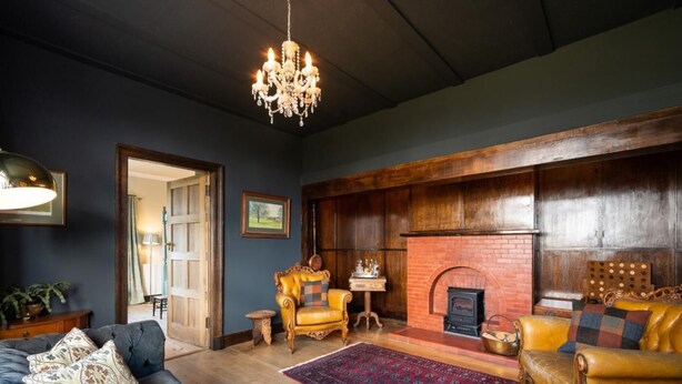 Een woonkamer met een donkerblauwe bank, een gele fauteuil, een open haard en een kroonluchter.