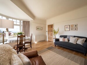 A living room with a sofa, a chair, a table, and a lamp. There is a window with curtains, a radiator, and a door.
