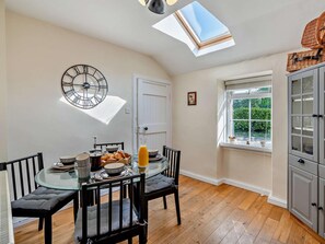 A dining area with a round glass table, black chairs, a wall clock, and a window with a view of greenery.