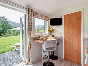 Ein Zimmer mit Holztür, einem an der Wand befestigten Fernseher, einem Fenster mit Vorhängen, einem kleinen Tisch mit Blumenvase und einem Stuhl.