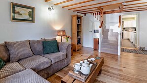 Sala de estar con un sofá, una mesa de centro con teteras, una escalera y una cocina visible a través de un puerta abierta.