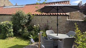 Un patio con muebles de mimbre y una mesa, rodeado de un jardín con diversas plantas y un muro de piedra.