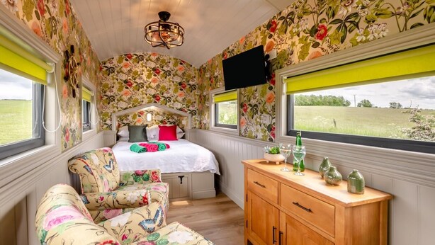Ett rum med blommig tapet, en säng med färgglatt sänglinne, ett träskåp och en tv på väggen.