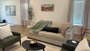Ett vardagsrum med en soffa, två fåtöljer, ett soffbord, en tavla och ett fönster med gardiner.