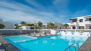 Una piscina con bordo in legno, circondata da edifici bianchi con balconi e palme.