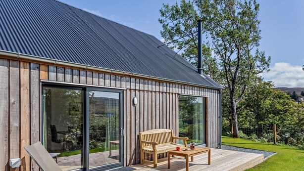 A modern building with a wooden exterior, large glass doors, a wooden deck with outdoor furniture, and a grassy garden.