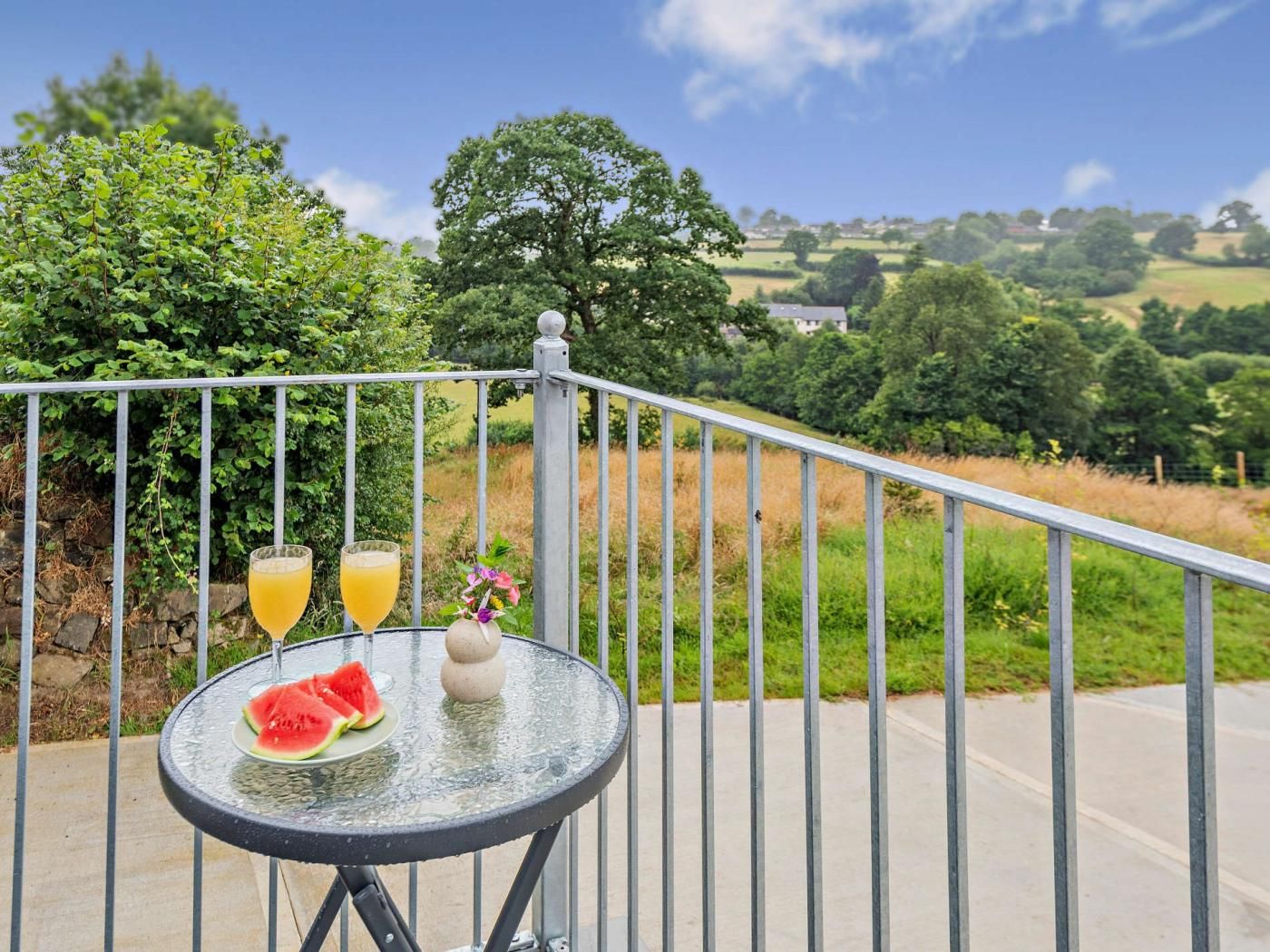 A balcony with a table, a glass of orange juice, a slice of watermelon, and a decorative vase.