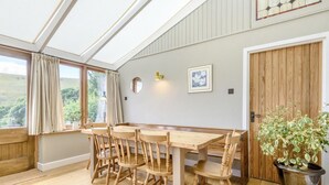 A dining area with a wooden table and chairs, a potted plant, and a view of the outdoors through large windows. - 3 Bed in Buxton (oc-96211) (Cressbrook)