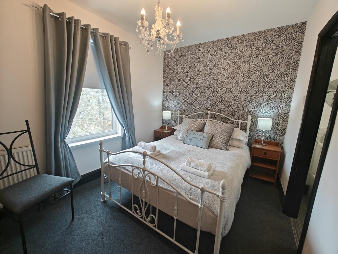 Ein Schlafzimmer mit einem Bett, einem Sessel, einem Fenster mit Vorhängen, einer Chandelüster und einer Wand mit Muster.