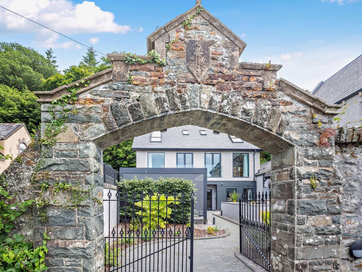 A stone archway leading to a modern house with a paved driveway and a metal gate.