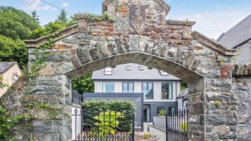 A stone archway leading to a modern house with a paved driveway and a metal gate.