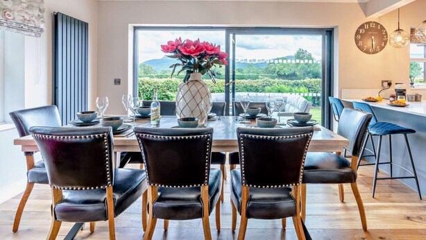 A dining area with a large table, four chairs, a vase with flowers, and a view of greenery outside. - 4 Bed in Middlesbrough (oc-96213) (Great Broughton)