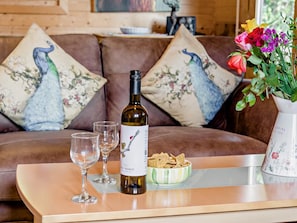 Ein gemütliches Wohnzimmer mit braunem Sofa, dekorativen Kissen, einer Weinflasche, zwei Gläsern und einer Schale mit Snacks auf einem Holztisch.