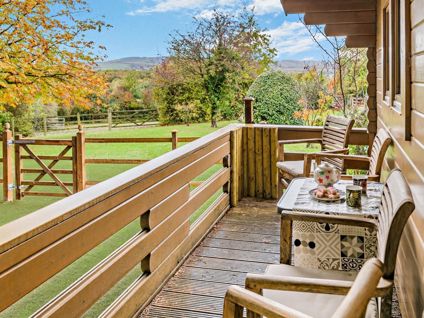 Une terrasse en bois avec une table et des chaises, donnant sur une zone herbeuse agrémentée d’arbres et bordée d’une clôture.
