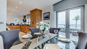 A modern dining area with a glass table, grey upholstered chairs, and a view of the ocean through large windows. - 2 Bed in Tenby (oc-96239) (Tenby)