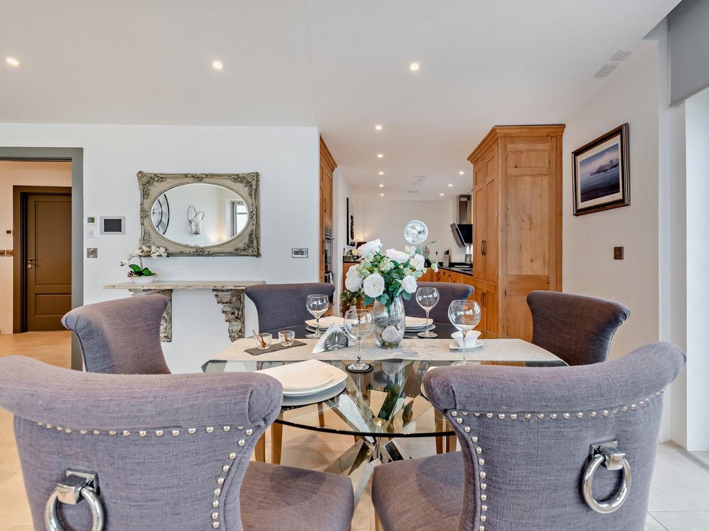 Un coin repas avec une table ronde, quatre chaises rembourrées, un vase de fleurs et un grand miroir au mur.