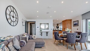 A modern living room with a sectional sofa, a dining area with chairs, a kitchen in the background, and a large wall clock. - 2 Bed in Tenby (oc-96239) (Tenby)