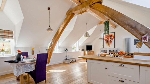 A spacious loft with a high ceiling, exposed wooden beams, a dining area with a purple chair, a kitchen with white cabinets, and a large window. - 1 Bed in Inverkeithing (oc-96221) (Inverkeithing)