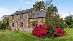 Una casa in pietra con tetto spiovente, una porta di legno e un cortile recintato con cespugli fioriti di colore rosso.