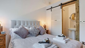 Een net opgemaakt bed met kussens, een nachtkastje met een lamp, een houten nachtkastje, en een badkamer met een wastafel en spiegel.