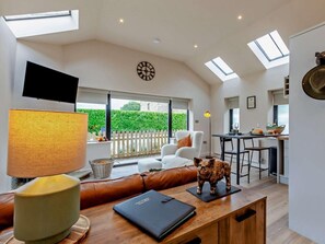 Uma sala de estar moderna com um sofá, uma mesa com abajur, uma televisão e vista para um jardim através de grandes janelas.