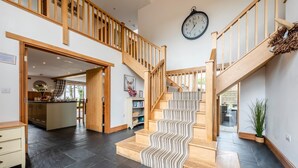 A spacious interior with a central staircase, wooden railings, a kitchen area, and a dining space. - 6 Bed in Holsworthy (oc-lympb) (Bradworthy)