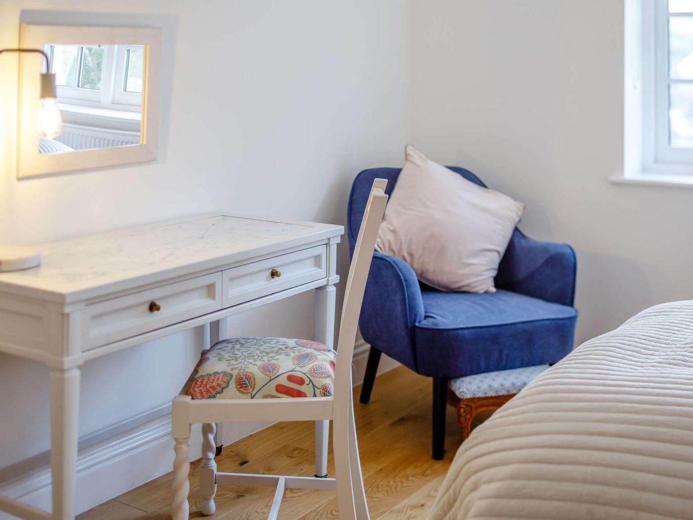 Une chambre avec une commode blanche, une chaise bleue avec un coussin et un lit recouvert d’une couette blanche.