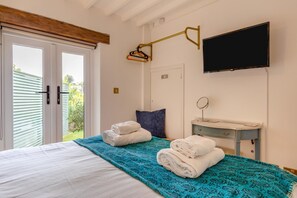 Ein Schlafzimmer mit einem Bett, einem Nachttisch mit Spiegel, einem an der Wand befestigten Fernseher und einem Fenster mit Blick auf Grünflächen.