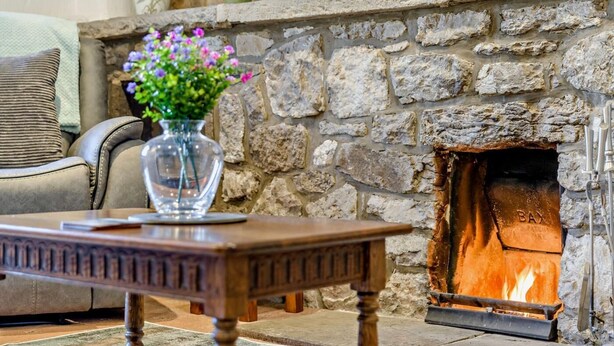 A stone fireplace with a fire, a wooden table in front, and a sofa with a cushion. - 3 Bed in Carnforth (oc-96319) (Yealand Redmayne)
