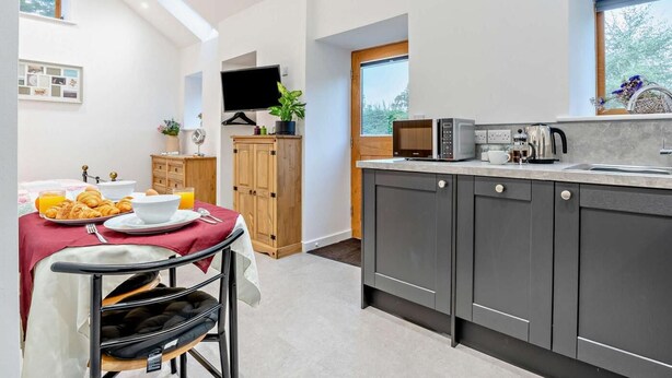 A modern kitchen with a dining area, a TV mounted on the wall, and a window with a view of trees. - 1 Bed in Pickering (oc-96309) (Allerston)