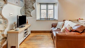 A cozy living room with a stone wall, a television on a wooden stand, a window with blinds, a brown leather sofa, and a white cabinet. - 1 Bed in Launceston (oc-96333) (Saint Giles on the Heath)