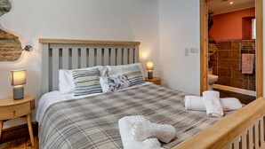 A bedroom with a wooden headboard, a plaid bedspread, two bedside lamps, and a stone wall. - 1 Bed in Launceston (oc-96333) (Saint Giles on the Heath)