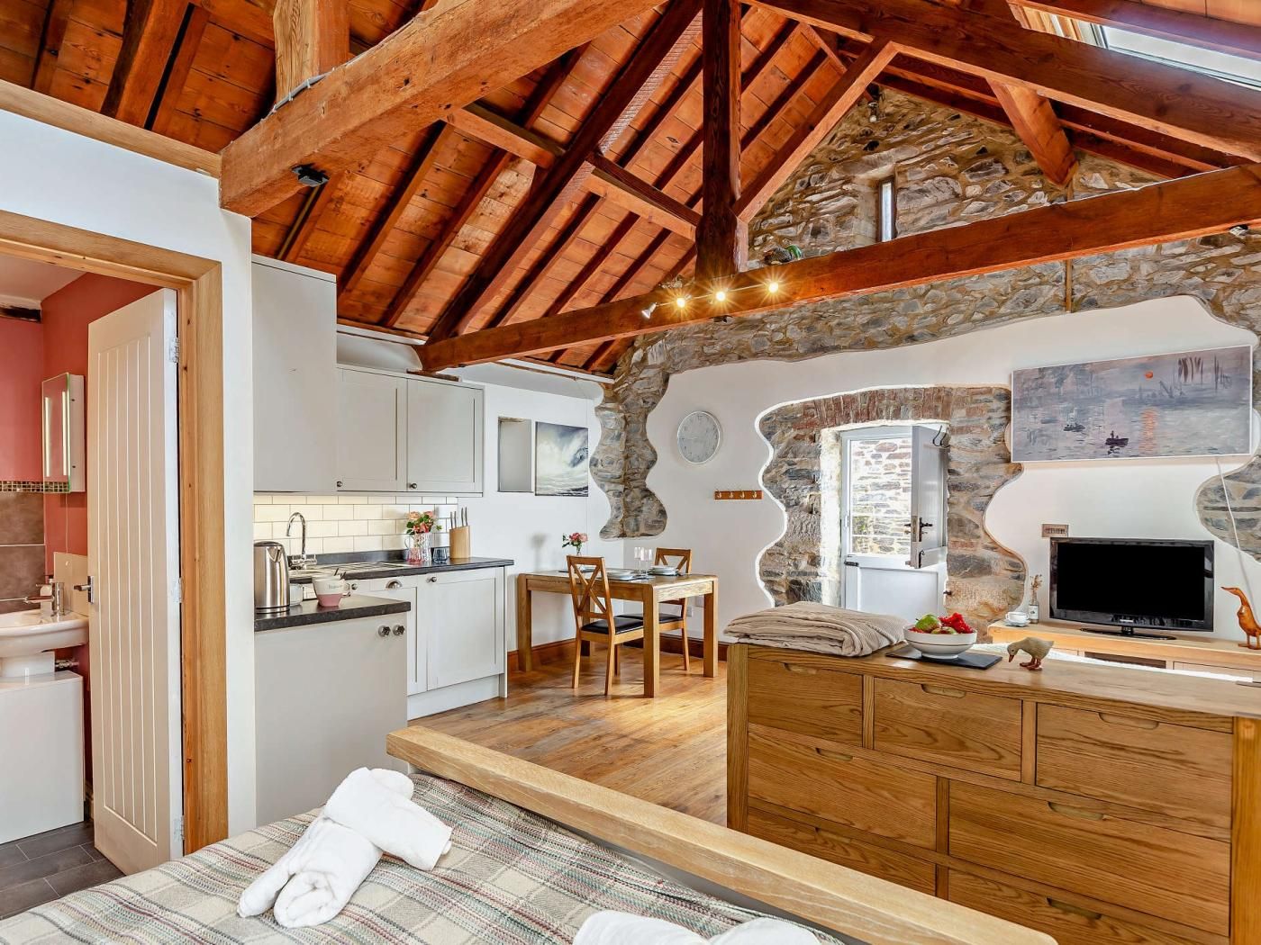A rustic interior with exposed wooden beams, a stone wall, a kitchen with white cabinets, and a dining area with a wooden table and chairs.