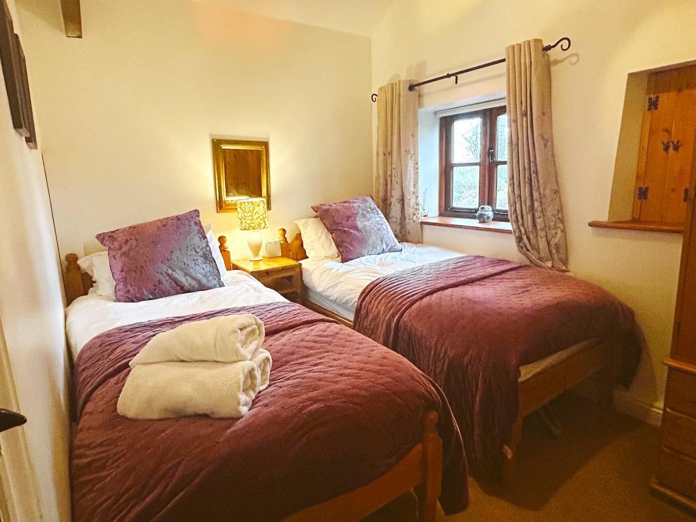 A room with two beds, each with a headboard, a bedside table with a lamp, and a window with curtains.