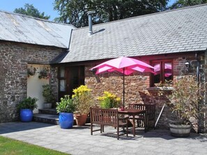 A stone building with a tiled roof, a patio with a wooden table and chairs, and potted plants. - 2 Bed in Holsworthy (oc-beeba) (Thornbury)