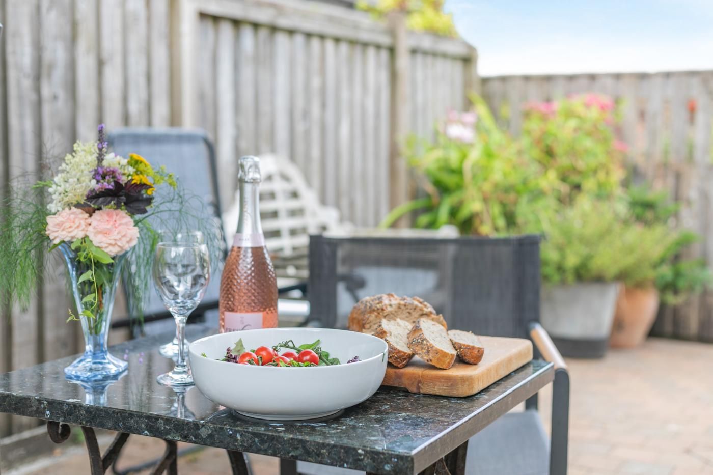 An outdoor dining table with a bowl of salad, a bottle of rosé wine, and slices of bread on a wooden board.