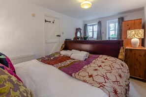 A bedroom with a bed, a wooden headboard, a nightstand with a lamp, a window with curtains, and a door with a handle. - 1 Bed in Holsworthy (oc-dairc) (Thornbury)