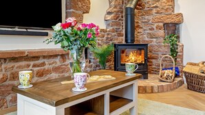 A stone fireplace with a TV mounted above it, a wooden table with a vase of flowers, and a basket of firewood. - 2 Bed in Carlisle (oc-96302) (Scaleby)