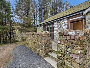 A stone building with a wooden door and a window, surrounded by a stone wall and trees.
