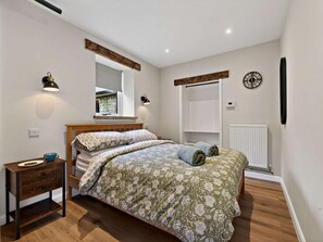A bedroom with a wooden bed, a bedside table, a nightstand, a window, a wall-mounted clock, and a radiator.