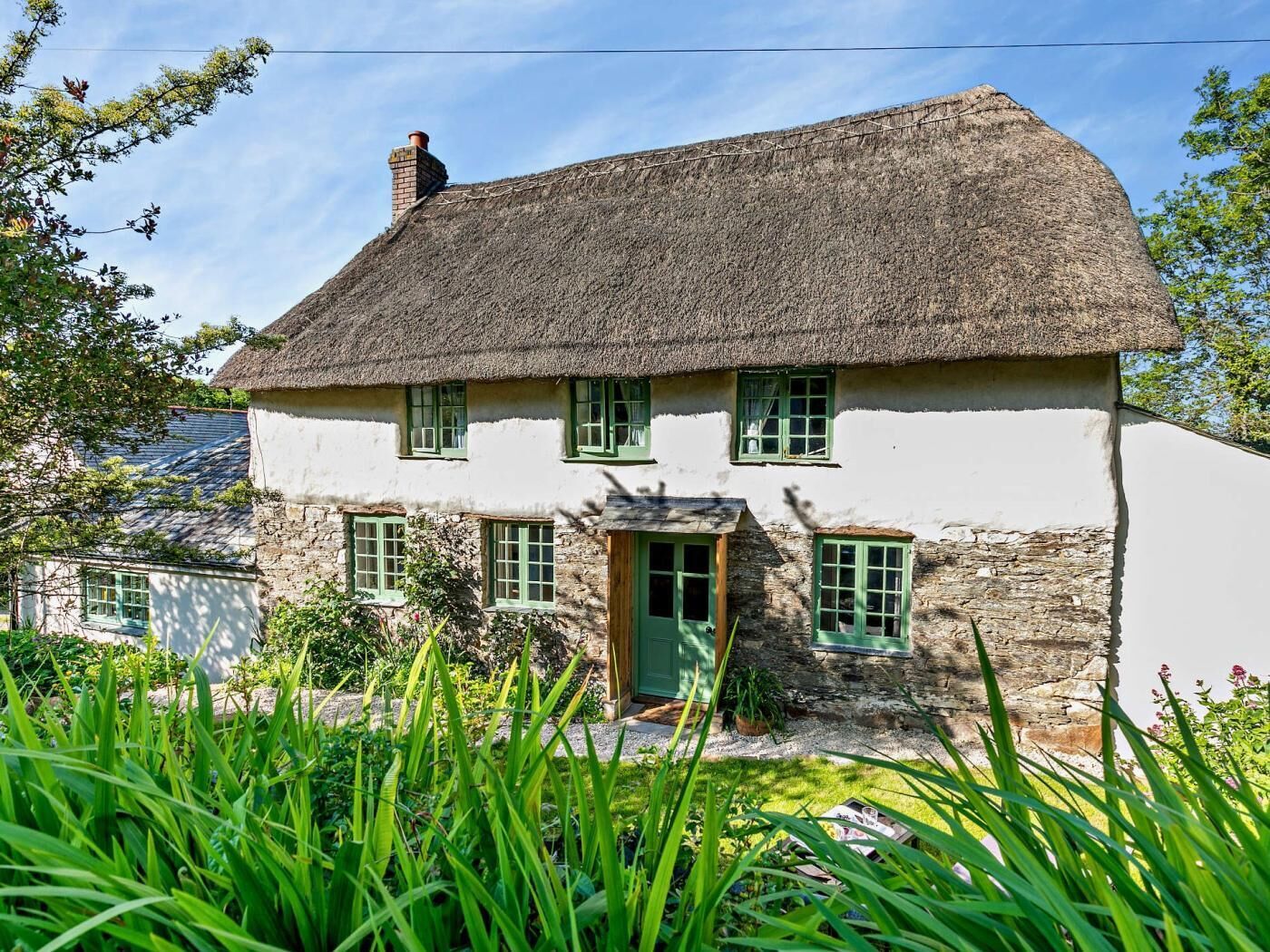 A traditional thatched-roof house with a stone foundation, green windows, and a small front porch.