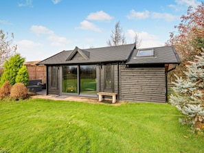 A modern log cabin with a glass door entrance, a small patio with a bench, and a well-maintained lawn. - 1 Bed in Maryport (oc-96373) (Dearham)