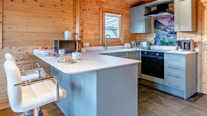 A wooden cabin interior with a kitchen featuring a central island, a dining area with a white chair, and a window with a view of the outdoors. - 2 Bed in Middlesbrough (oc-96344) (Skutterskelfe)