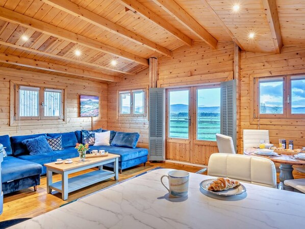 A cozy wooden cabin interior with a sofa, dining table, and large windows offering a view of the outdoors.