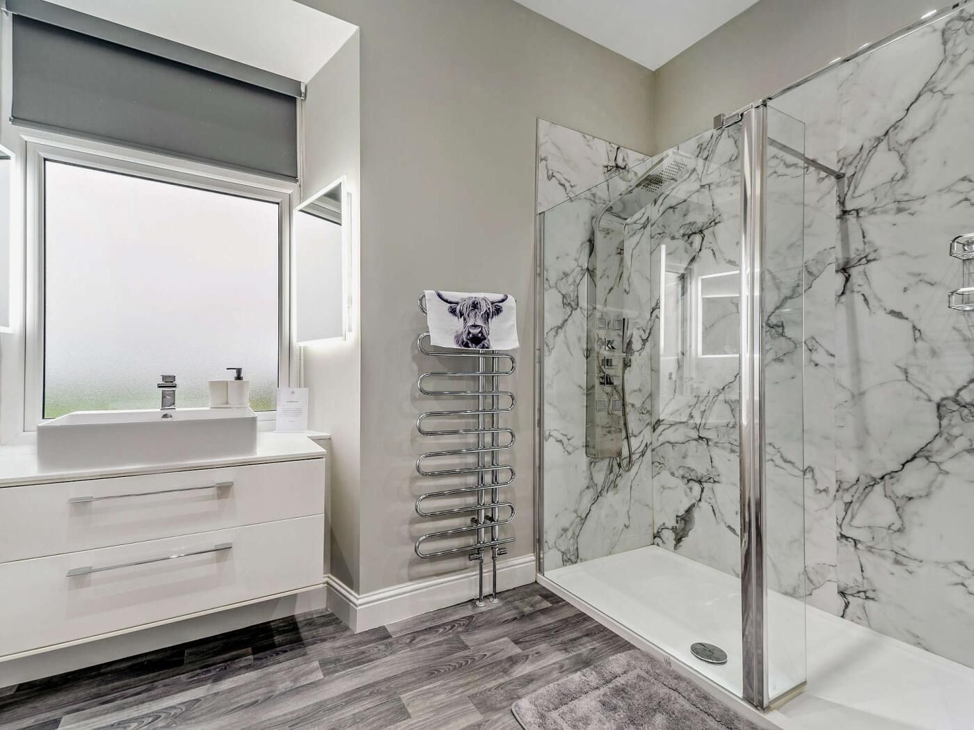 A modern bathroom with a marble shower, a white vanity with a window, and a towel rack with a towel.
