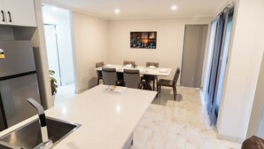 A modern kitchen with a central island, a dining area with a table and chairs, and a large window with curtains. - The Happy Place (Queens Park)