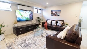 A living room with a TV, leather sofa, glass coffee table, and a patterned rug. - The Happy Place (Queens Park)