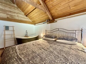 A bedroom with a wooden ceiling, a bed with patterned bedding, a metal-framed bed, a bedside table, and a built-in bathtub. - 1 Bed in Tiverton (oc-96405) (Washfield)